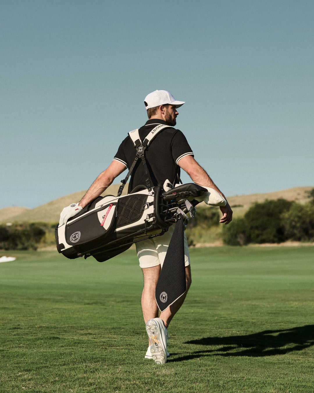 Lifestyle image of a male golfer walking across a golf course, viewed from behind, carrying a golf bag over his shoulder. He is wearing a white hat, white shorts, and a black polo shirt, with the course stretching out ahead of him.