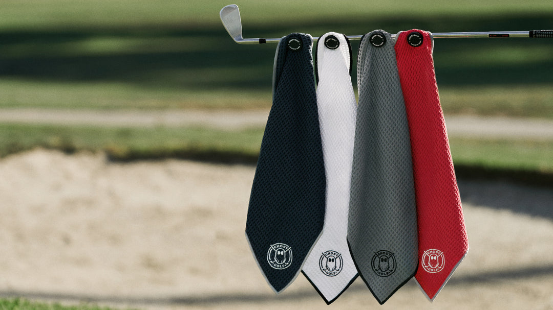 Lifestyle image of four Magnetic Greenside Towels in black, grey, white, and red hanging from a golf club near a greenside bunker. Each towel features a Ghost Golf emblem and branded magnetic attachment.