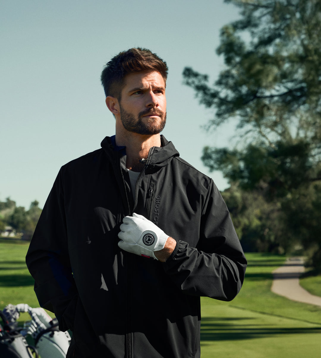 White male golfer on a golf course pulling down the zipper of his jacket while wearing a white golf glove.