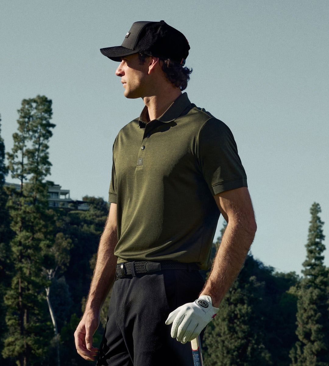 White male golfer on a golf course wearing a green polo tucked into black pants, a black belt, and a white golf glove, holding a golf club.