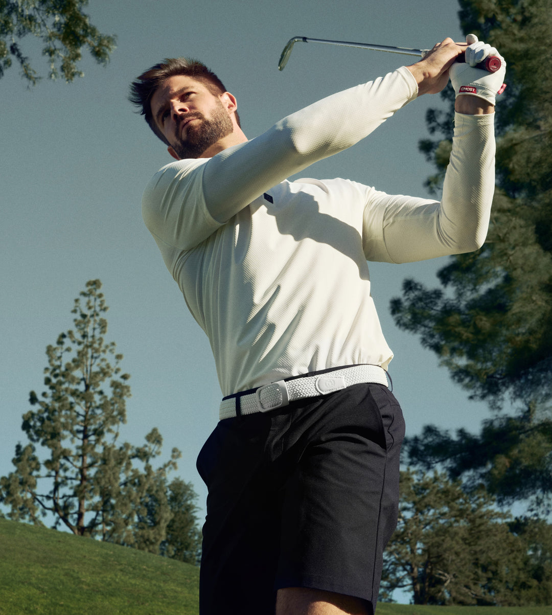 White male golfer captured mid-swing on a golf course, wearing a white long-sleeve polo paired with black shorts and a white belt.