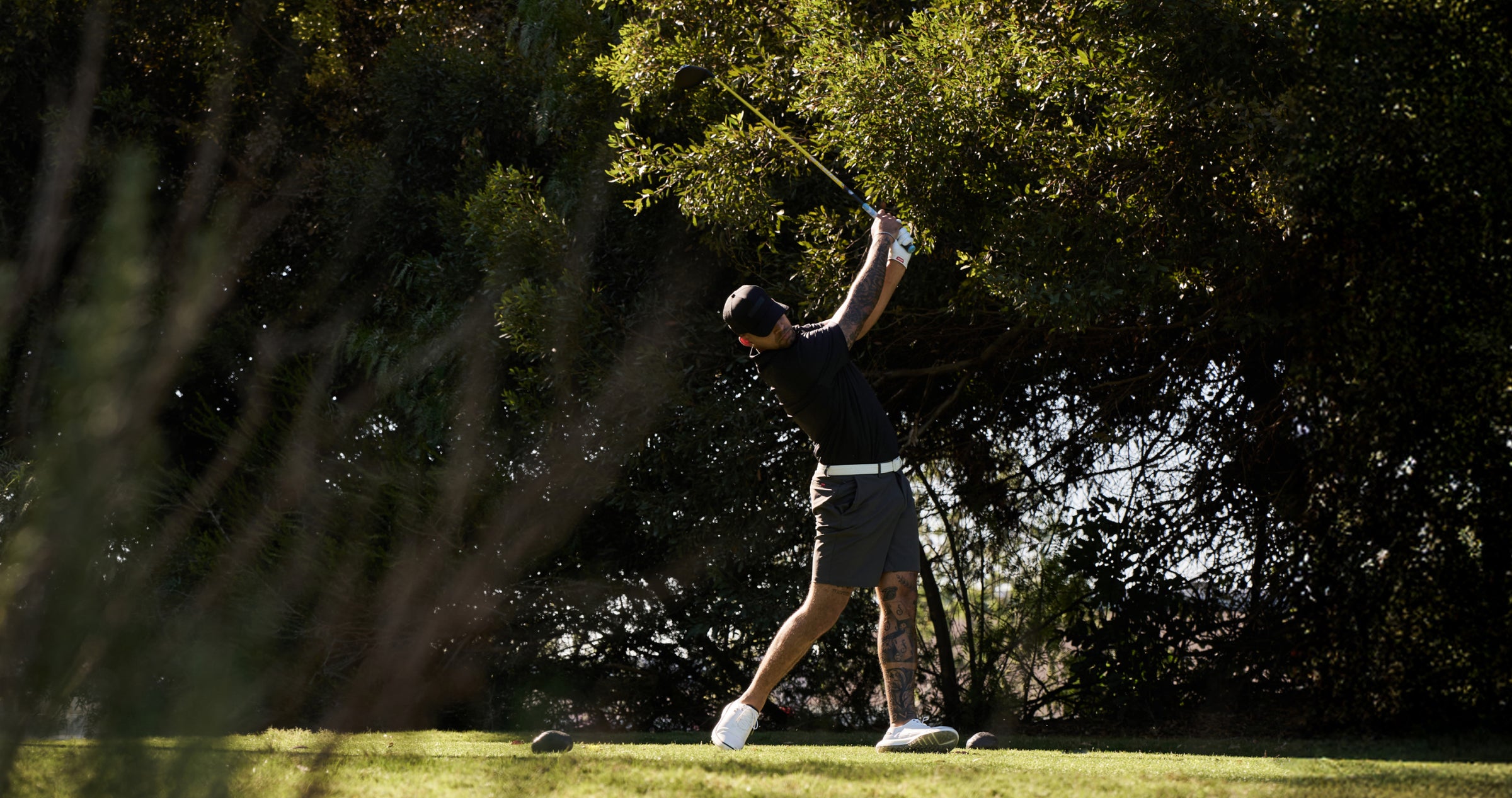 Male golfer mid-swing on a sunny golf course, wearing a black polo, white belt, and grey shorts