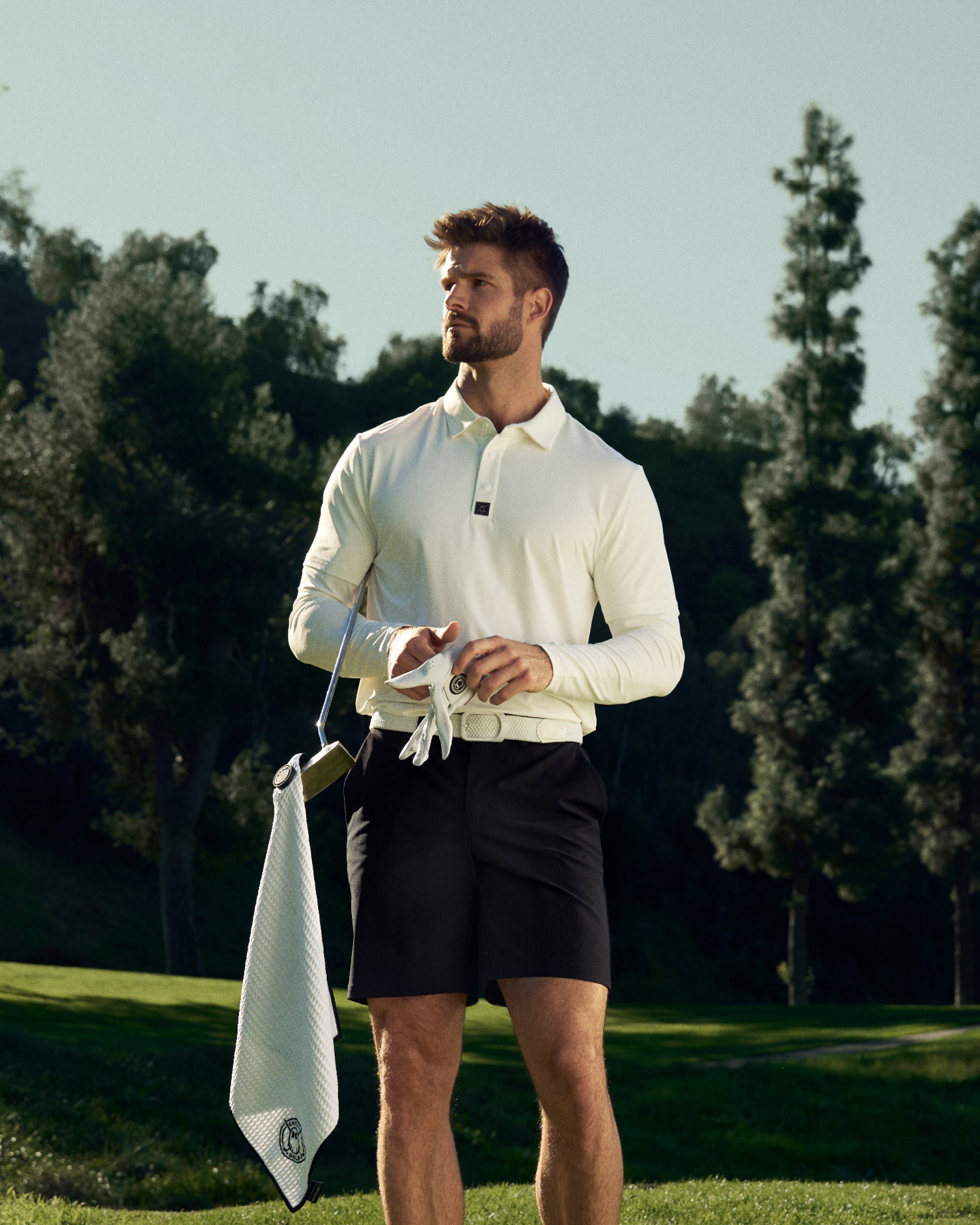 Golfer Standing on a Golf course Wearing a White Polo and Black Shorts. Golf Club and Towel in Hand.