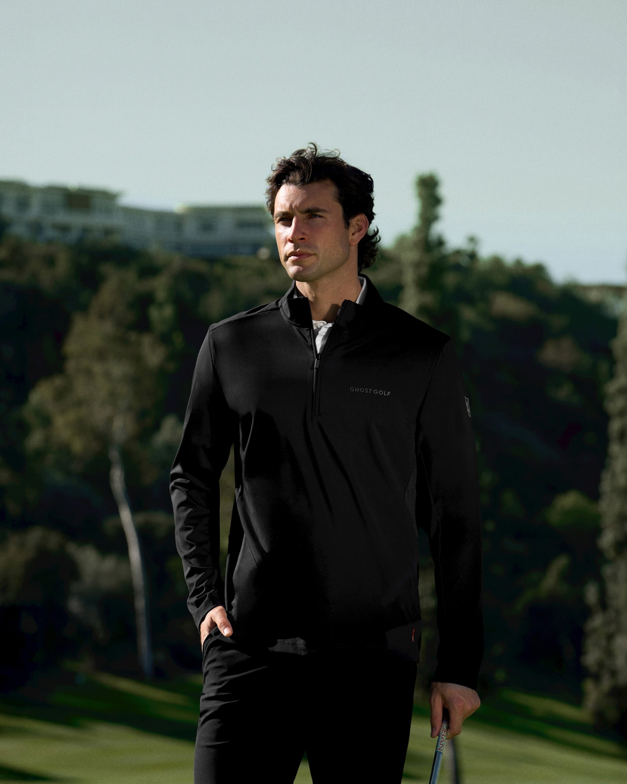 White male golfer on a golf course wearing a black quarter-zip mock jacket, hand in pocket.
