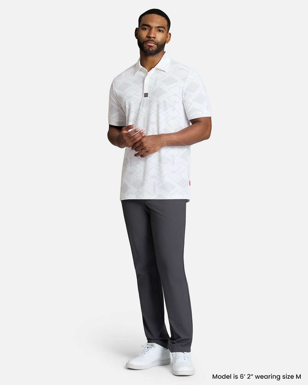 Full Body Front Shot of Black Male Model Wearing White Qualifier Argyle Polo with grey pants and white shoes. Polo is untucked. #color_white