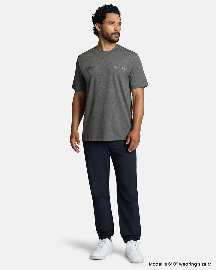 Full Body Front Shot of White Male Model Wearing Grey Qualifier Player Tee with navy pants and white shoes. #color_heather-grey