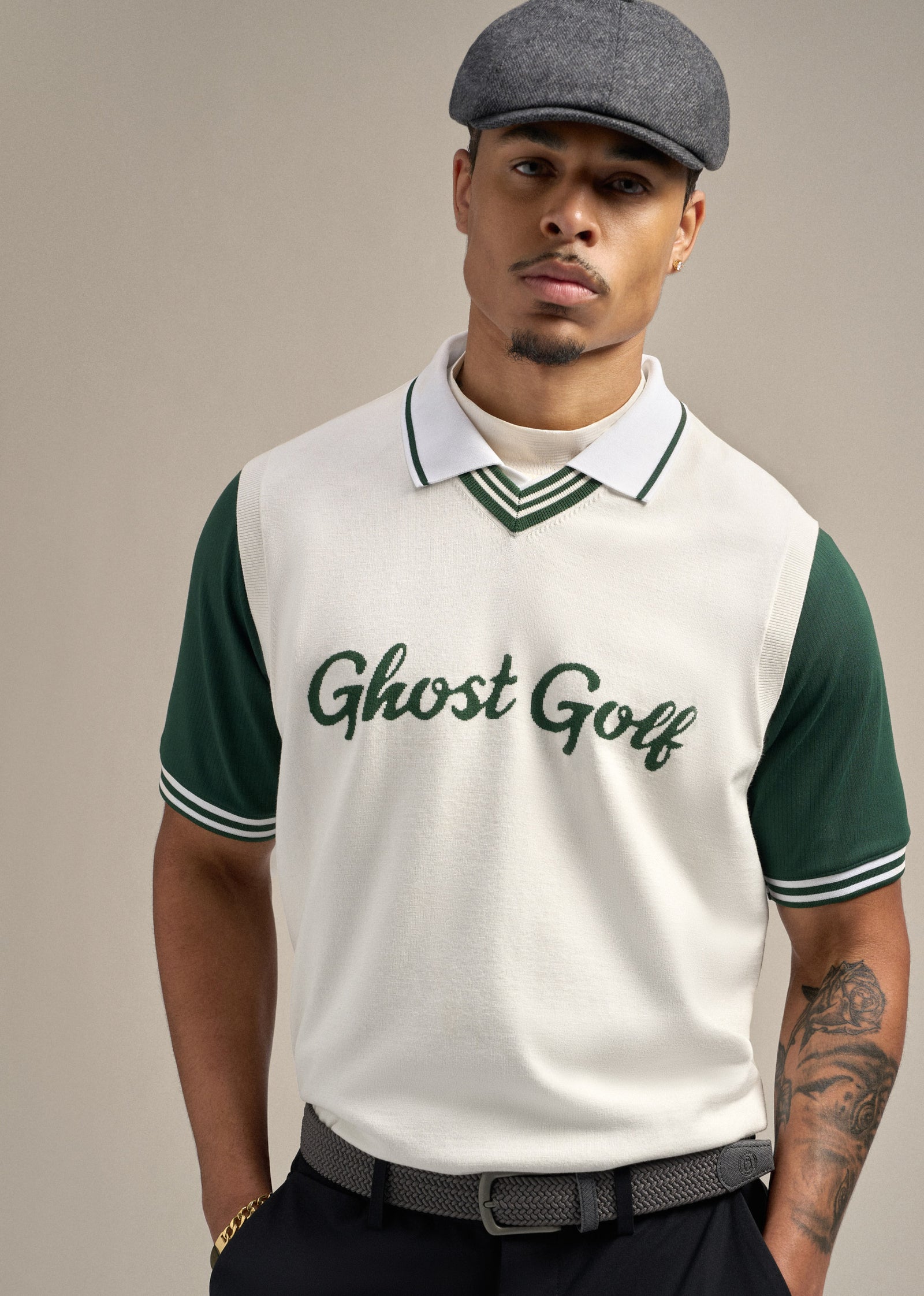 Front upper-body shot of a Black male model with his hands in the pockets of black pants, wearing a dark grey belt. He is dressed in a white sweater vest with a Ghost Golf embroidery over a white polo, paired with a flat cap. The model is looking straight ahead, creating a clean and confident pose.