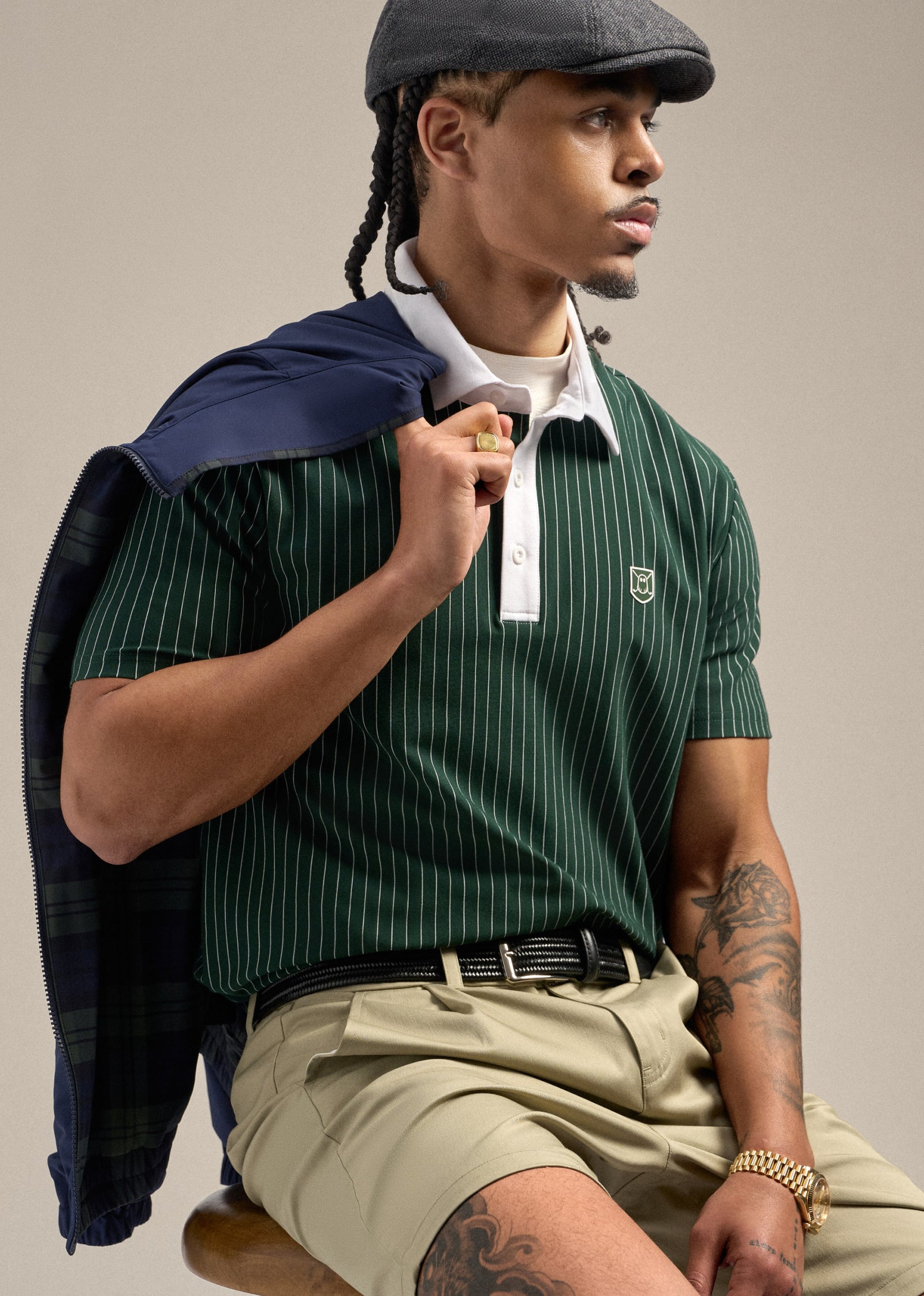 Upper body shot of a Black male model sitting on a chair and looking toward his left. He is wearing a green polo and a flat cap, with a jacket draped over his shoulder held by one finger. He is styled with sand wedge–colored shorts and a black belt, creating a relaxed yet refined golf-inspired look.