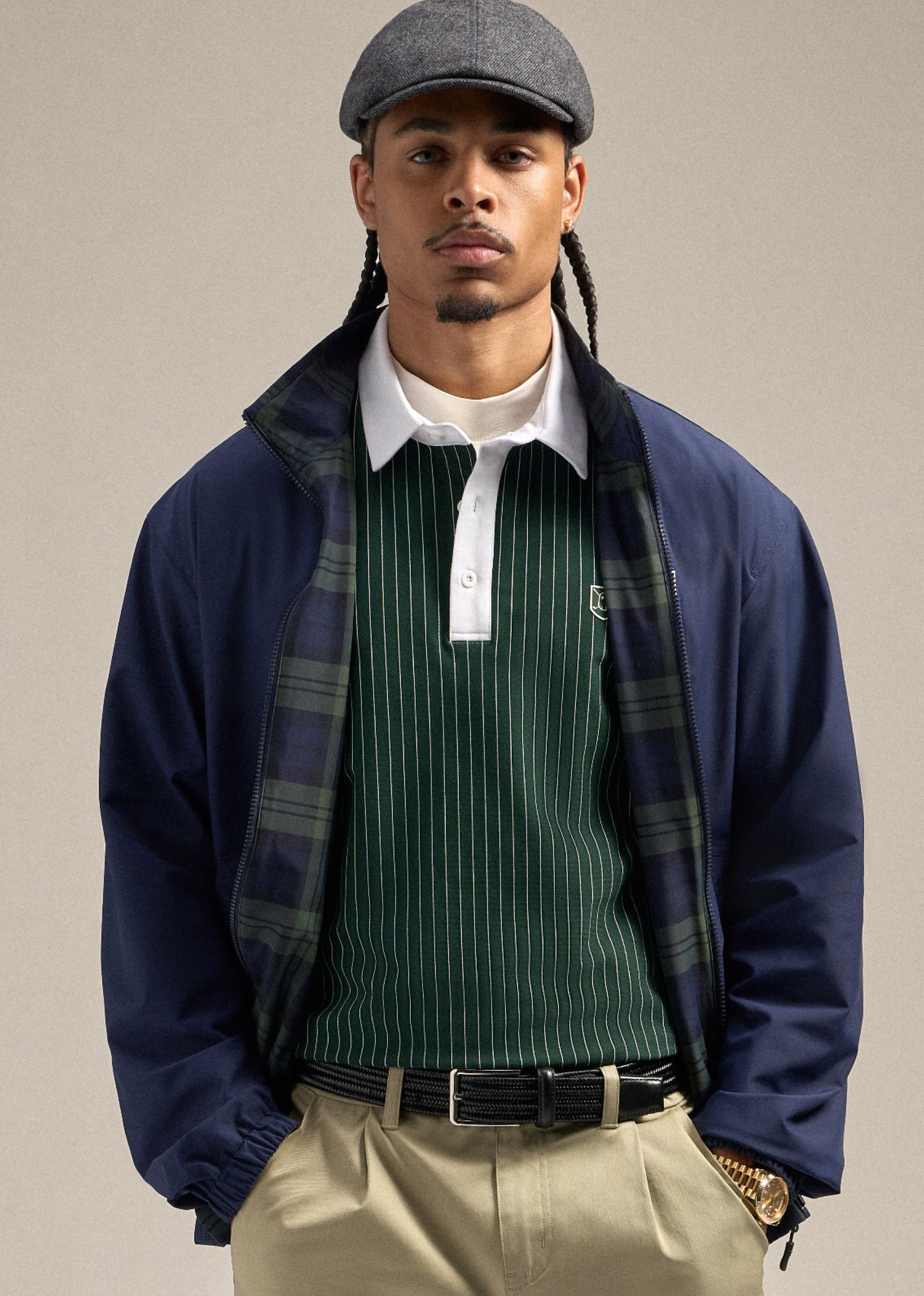 Black male model wearing a navy reversible spectator jacket with a green polo underneath. He is styled with a flat cap, sand wedge–colored shorts, and a brown belt. His hands are in his pockets, creating a relaxed and confident pose.