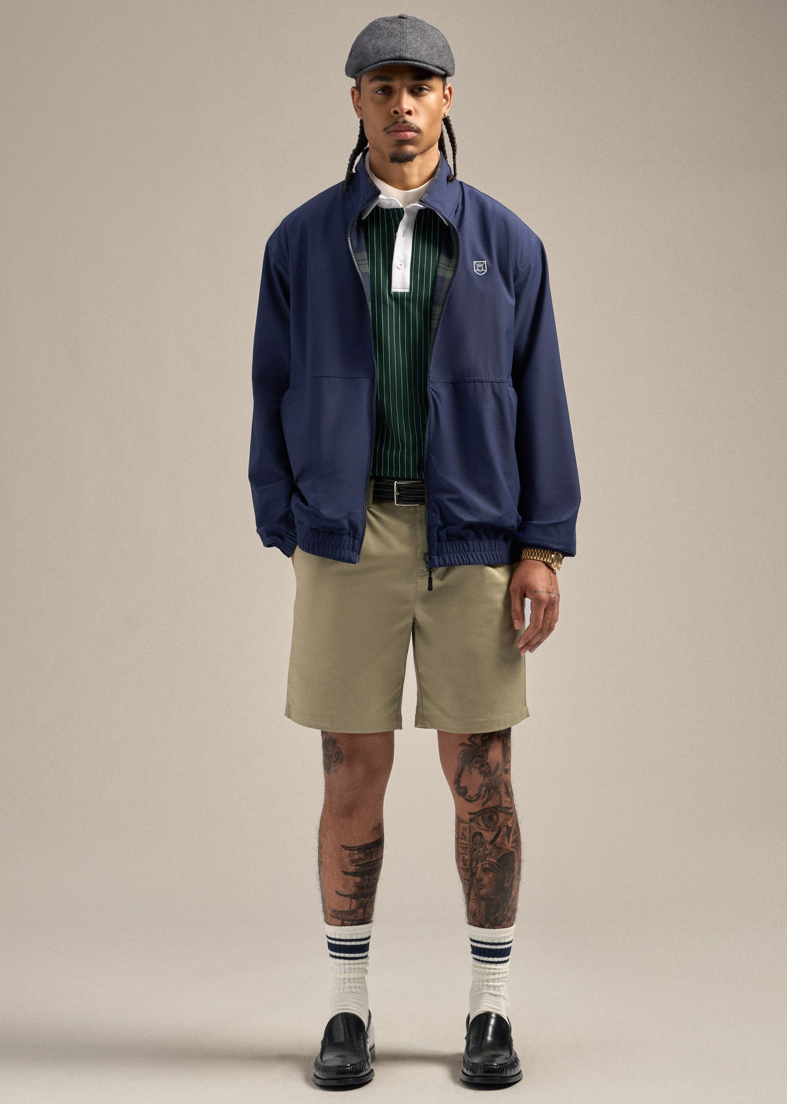 Full body shot of a Black male model wearing a navy reversible spectator jacket as a layer, with a green polo underneath. He is styled with a brown belt and sand wedge–colored shorts, paired with dress shoes and a flat cap. One hand is in his pocket while the other rests naturally at his side, looking straight ahead in a relaxed and confident pose.