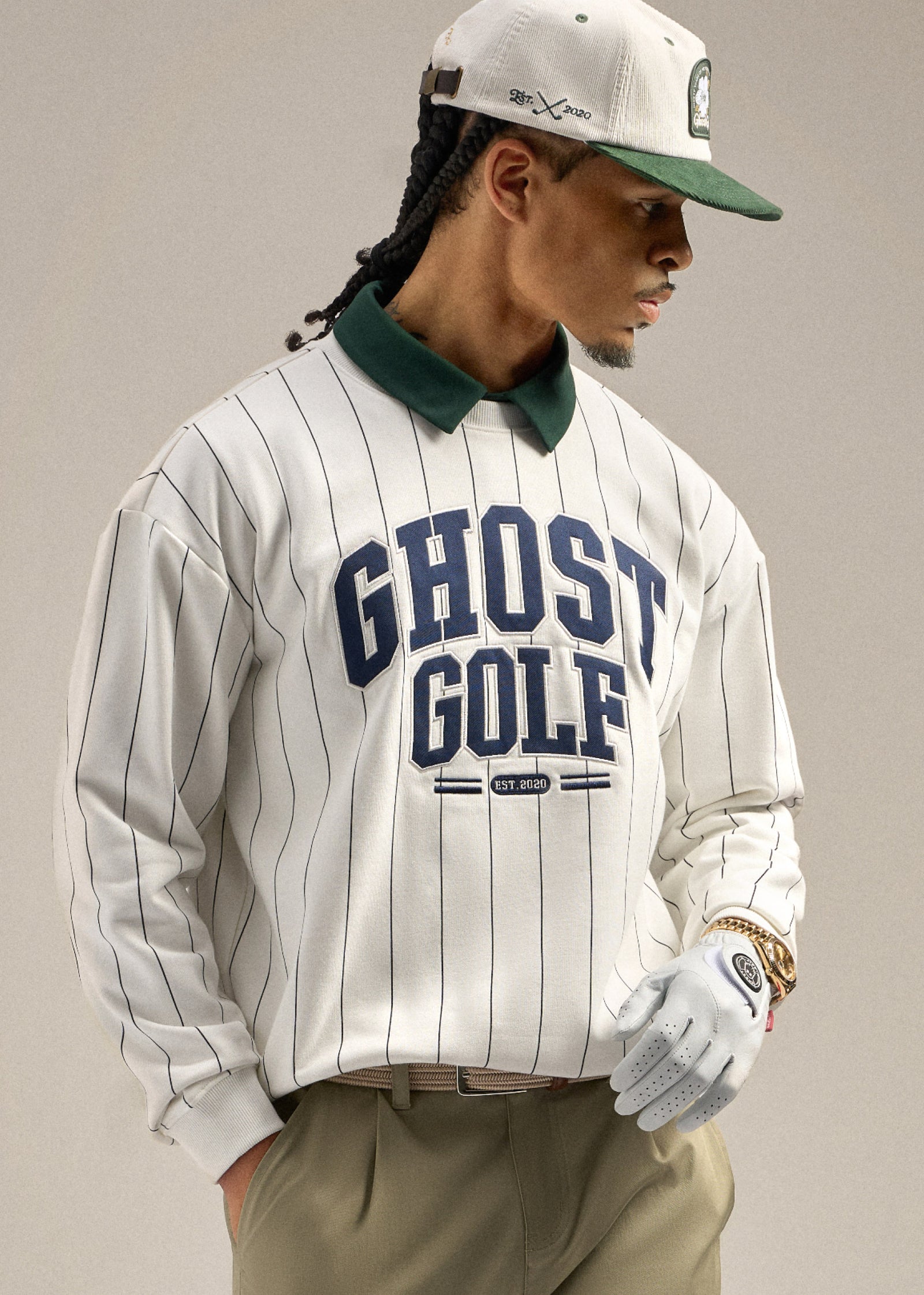 Black male model looking toward his right, wearing a white striped sweatshirt with Ghost Golf embroidered on it and a green polo underneath. He is styled with sand wedge–colored pants and a white cap, with one hand in his pocket, creating a relaxed and confident pose.