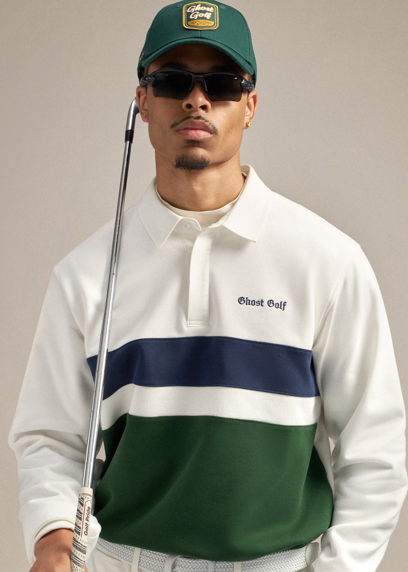 Close-up shot of a male model wearing a green hat and sunglasses. He is dressed in a white spectator sweatshirt polo and holding a golf club resting on his shoulder, looking straight ahead with a confident expression.