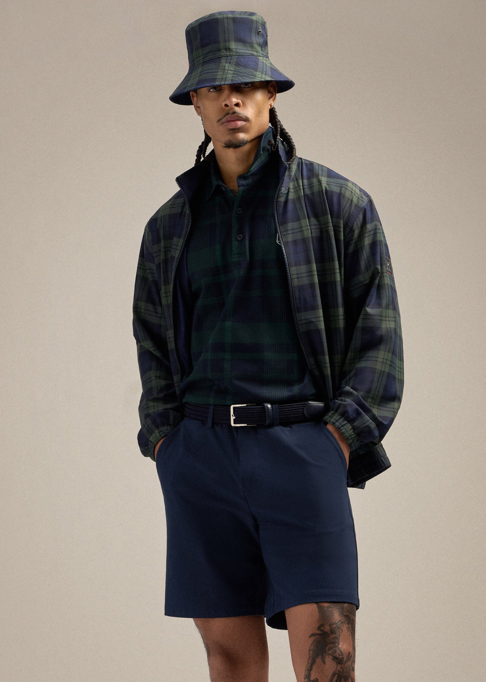 Black male model wearing a plaid polo, bucket hat, and jacket, styled with navy shorts and a navy belt. His hands are in his pockets as he looks straight ahead, creating a relaxed and confident pose.