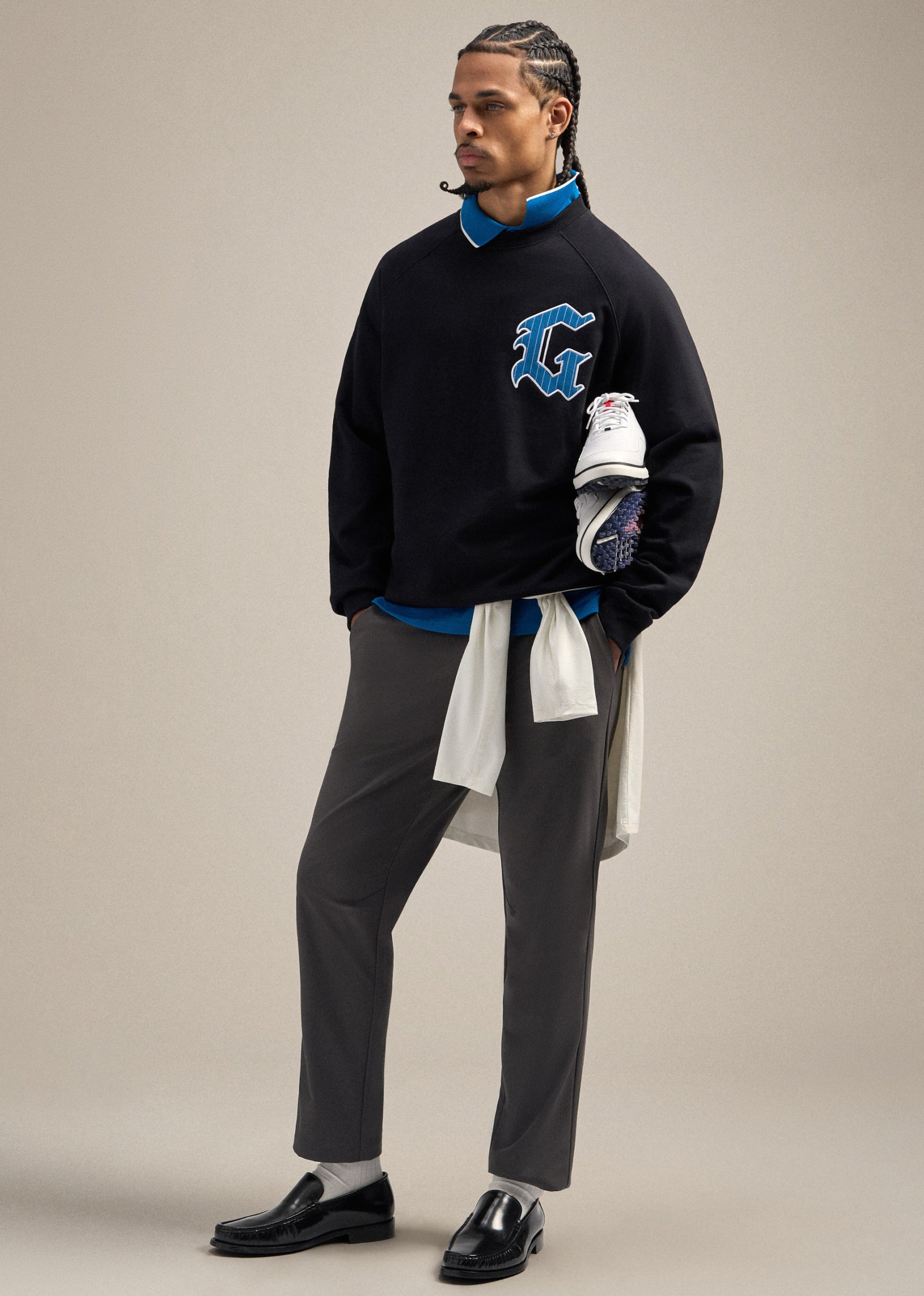 Black male model wearing a black sweatshirt with a large G hit on the left chest, layered over a royal blue polo. He is wearing grey pants and looking toward his left. Both hands are placed in the pockets of his pants, with golf shoes carried over his arm, creating a confident and styled golf-inspired pose.