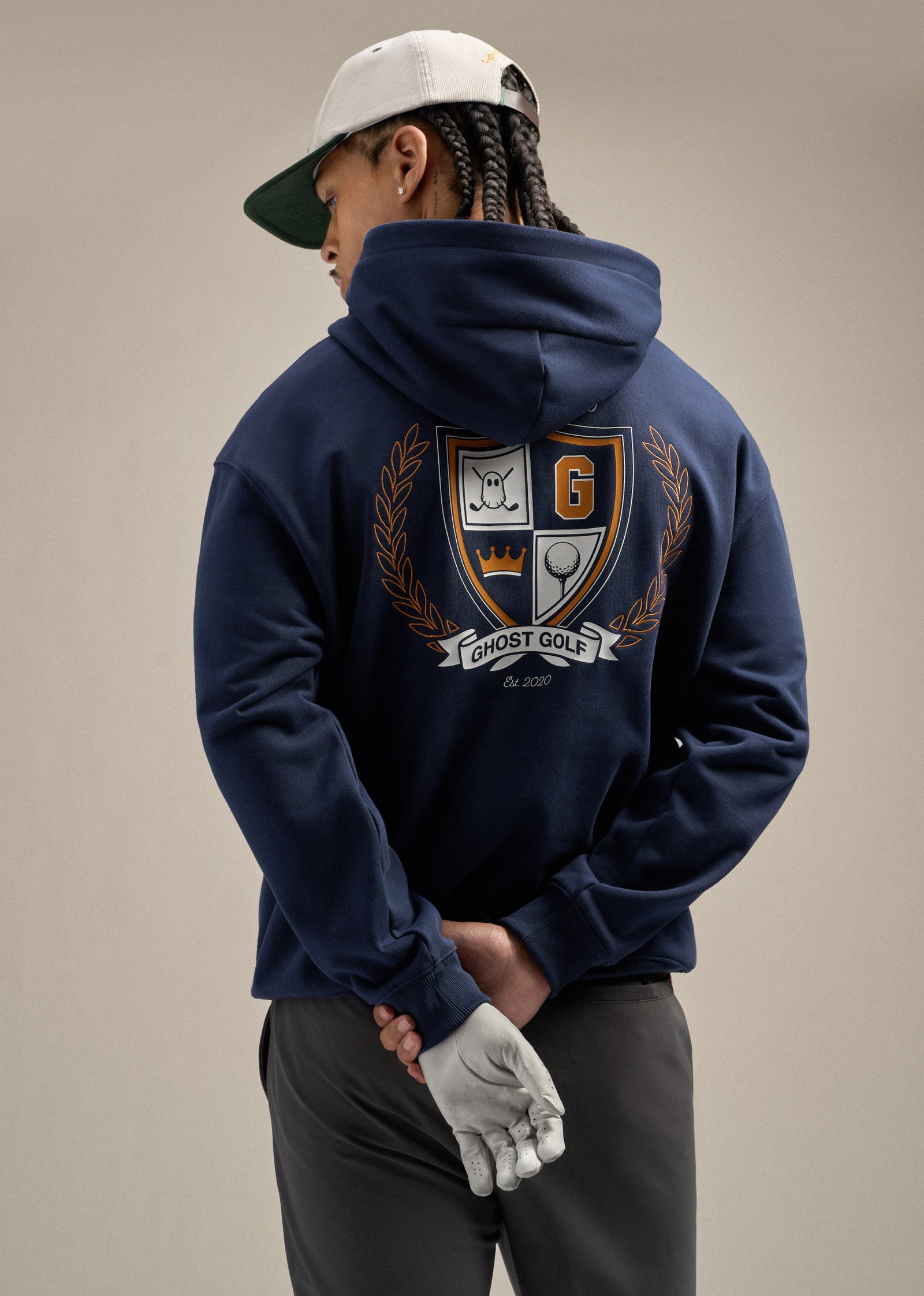 Black male model wearing a navy hoodie, grey pants, a white hat, and a white glove. The model is showcasing the back of the hoodie with a graphic hit, standing with his hands behind his back.