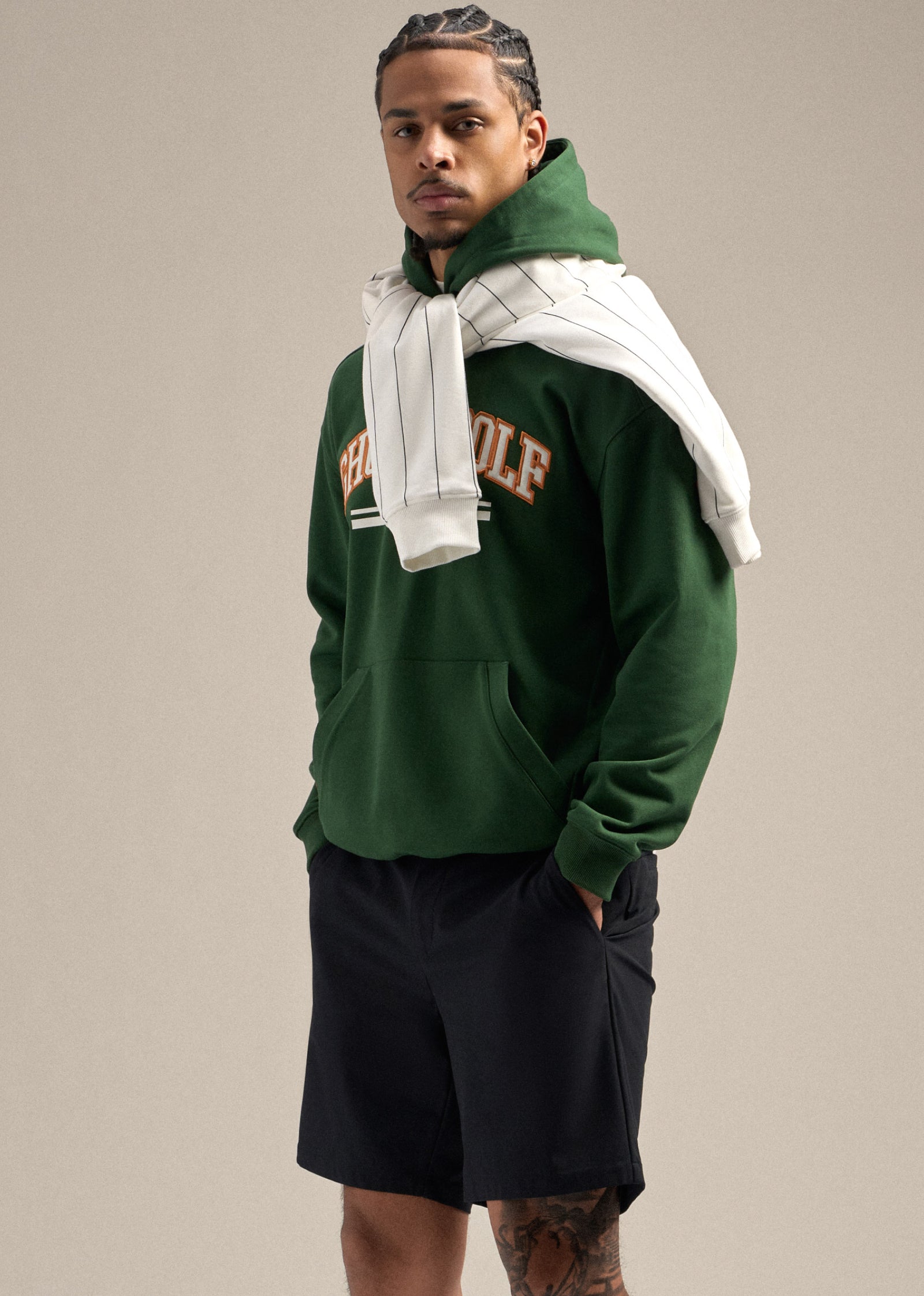 Black male model wearing a green hoodie with his hands in the pockets of black shorts. A white striped sweatshirt is draped around his neck, creating a relaxed and stylish look.