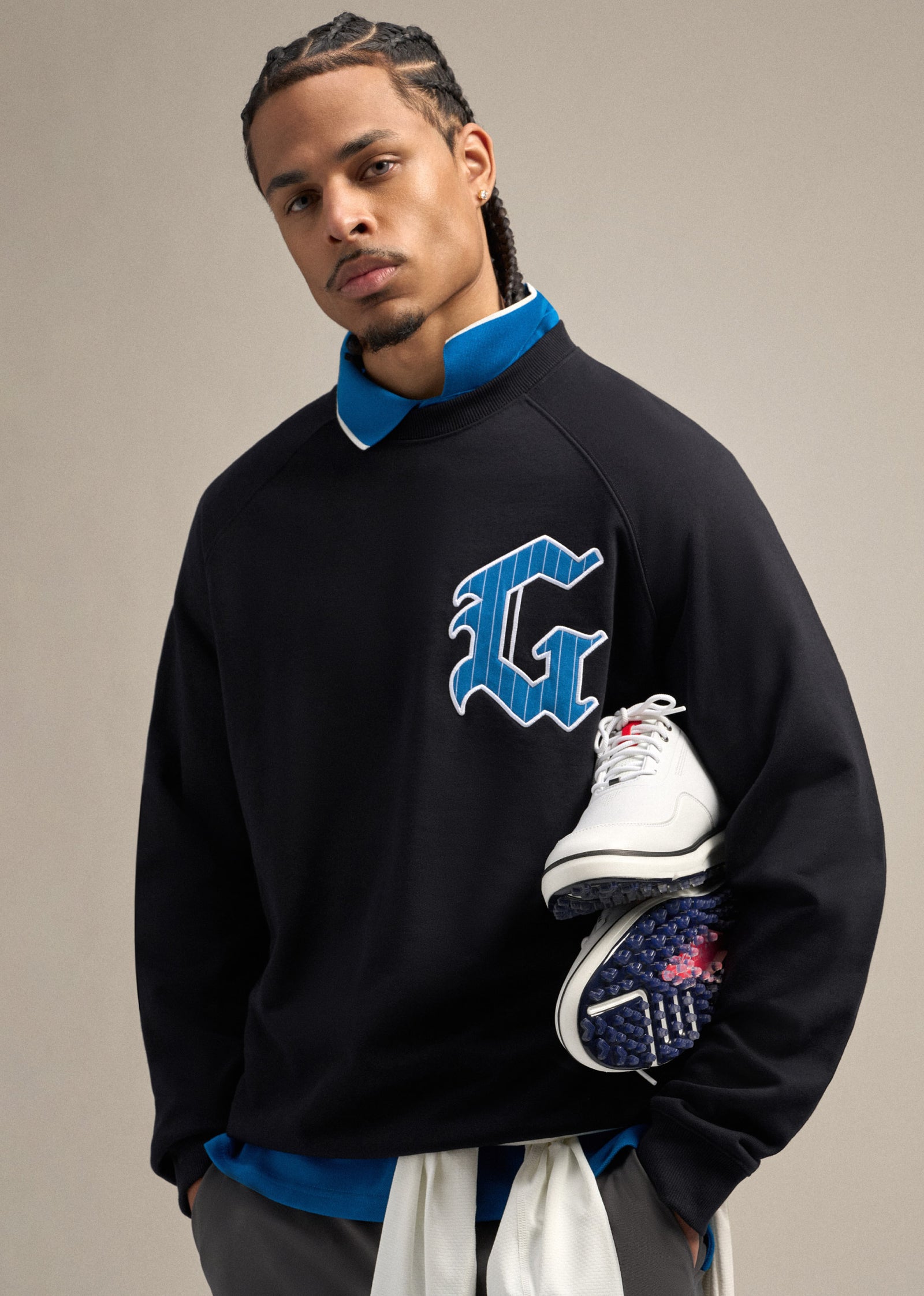 Black male model wearing a black sweatshirt with a large G hit on the left chest, layered over a royal blue polo. He is wearing grey pants and looking straight ahead. Both hands in pocket while one arm carries golf shoes, creating a confident and styled golf-inspired pose.
