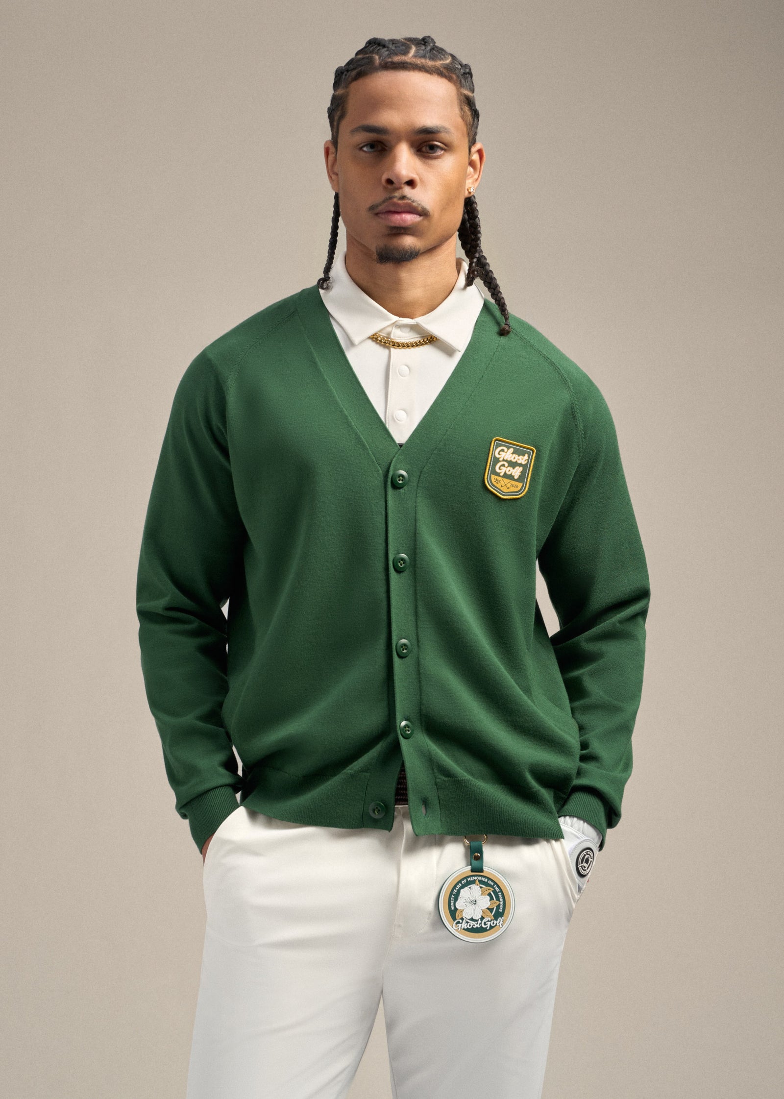 Full top shot of black male model wearing a green cardigan over a white polo. Both hands are in his pockets. He is wearing white pants with a Patrons Only golf accessory clipped to the front of the pants, creating a clean, golf-inspired look.