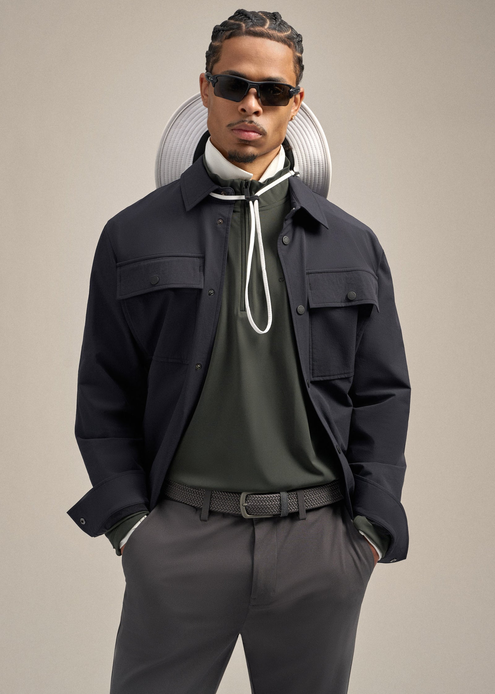 Black male model facing forward, wearing a black jacket layered over a green mock neck and a white polo underneath. His hands are in his pockets, styled with grey pants and a belt. A boonie hat is draped around his neck, hanging behind his head, creating a relaxed and modern look.