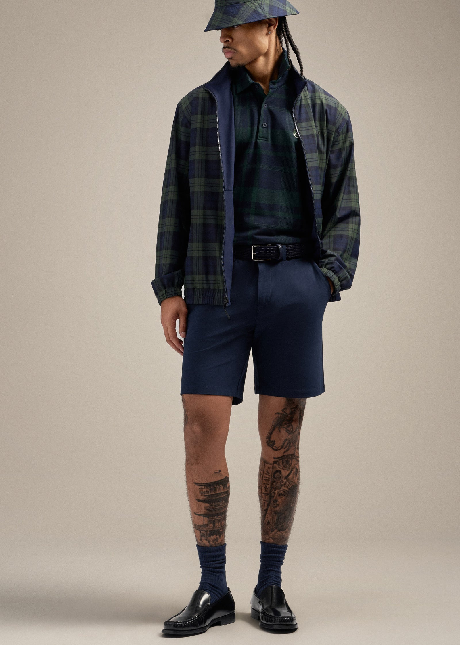 Black male model standing with one hand in his pocket, looking to his right. He is wearing a reversible plaid jacket, a bucket hat, a plaid polo shirt tucked into navy shorts, and a navy belt.