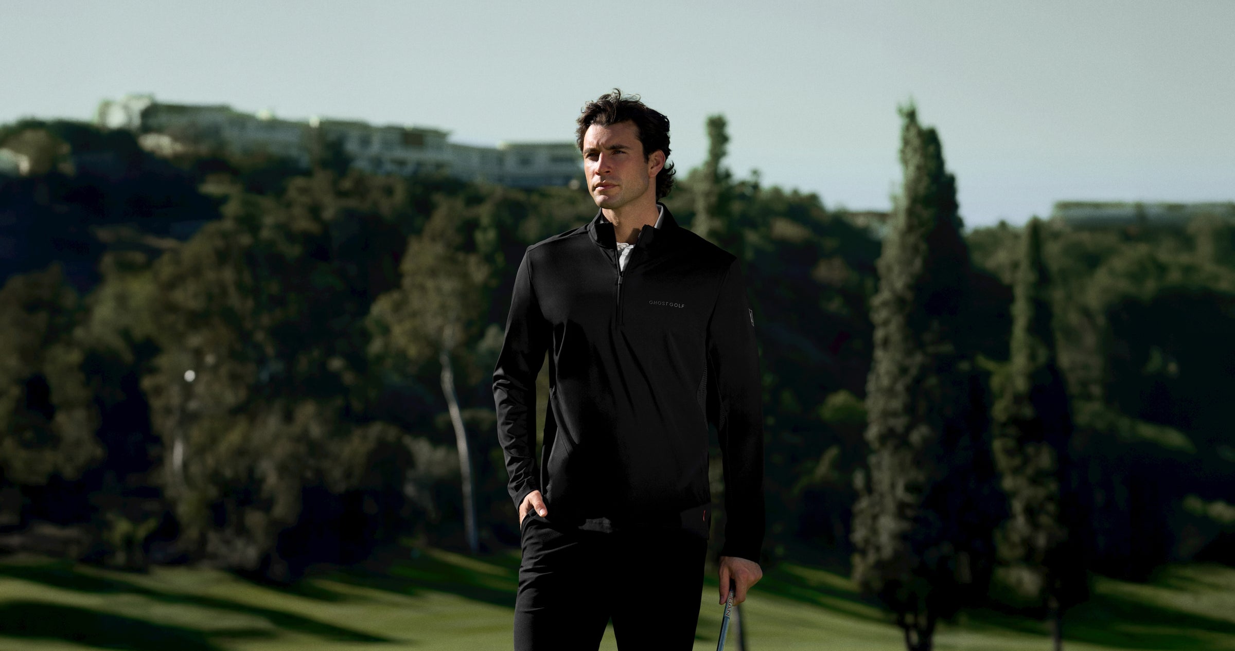 White male golfer on a golf course wearing a black quarter-zip mock jacket, hand in pocket.