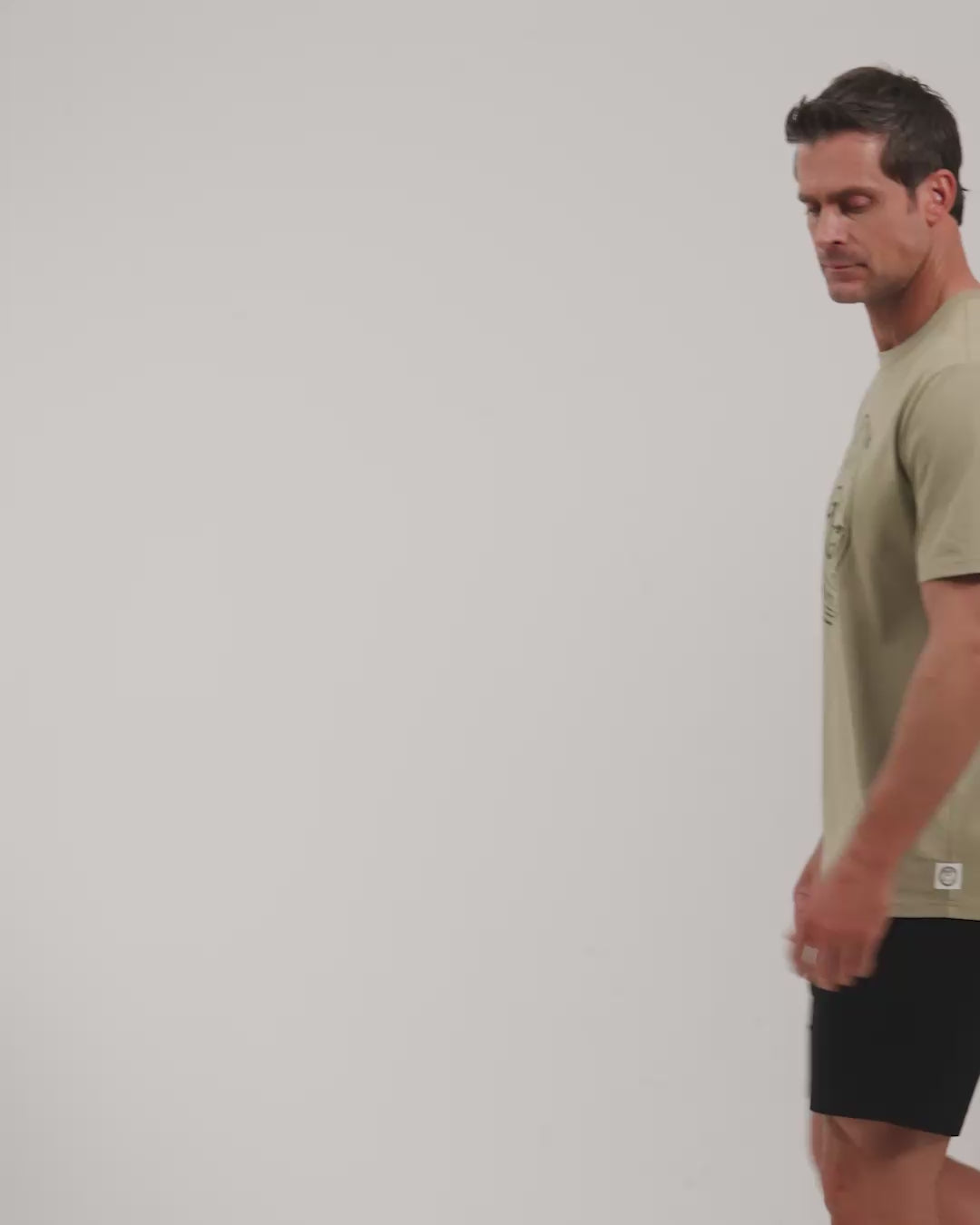 Male model walking naturally showcasing Sand Patrons Club Badge Tee from all angles wearing black shorts and white shoes #color_sand-wedge